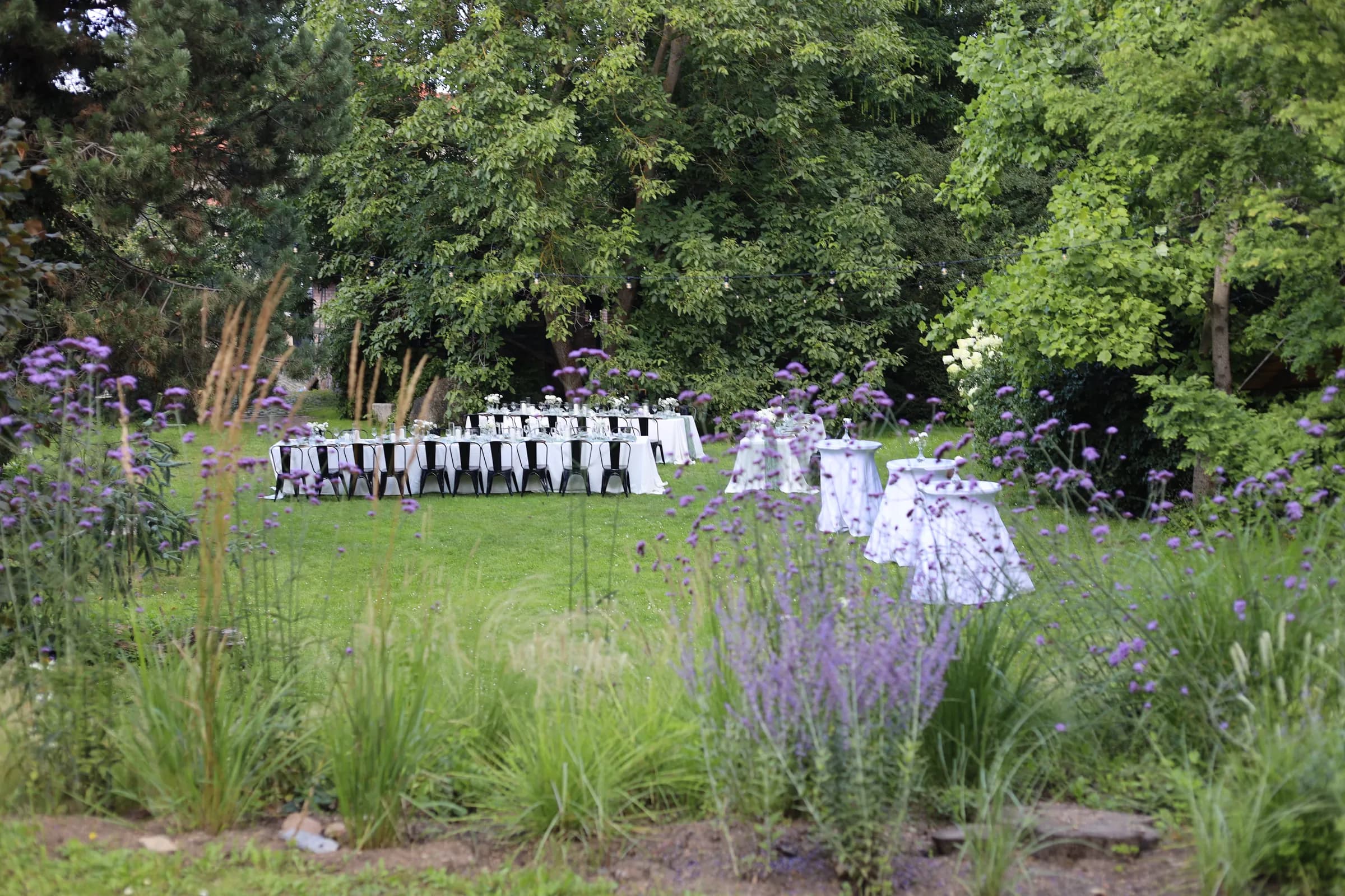 Mill garden with purple flowers and grasses in the flower bed, tables with white tablecloths, black chairs and white cocktail tables in the background