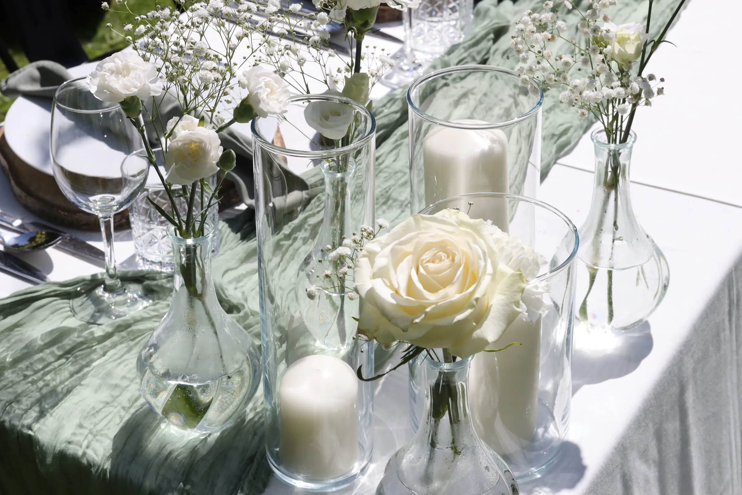 Carafe vases with a white rose, carnations and gypsophila between candles in glass cylinders on a sage gauze table runner