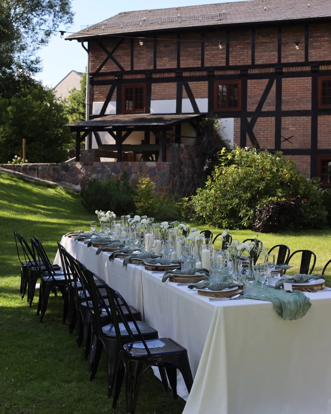 A long table for guests in front of a half-timbered building: white tablecloth, sage table runner, candles in glass and vases with flowers