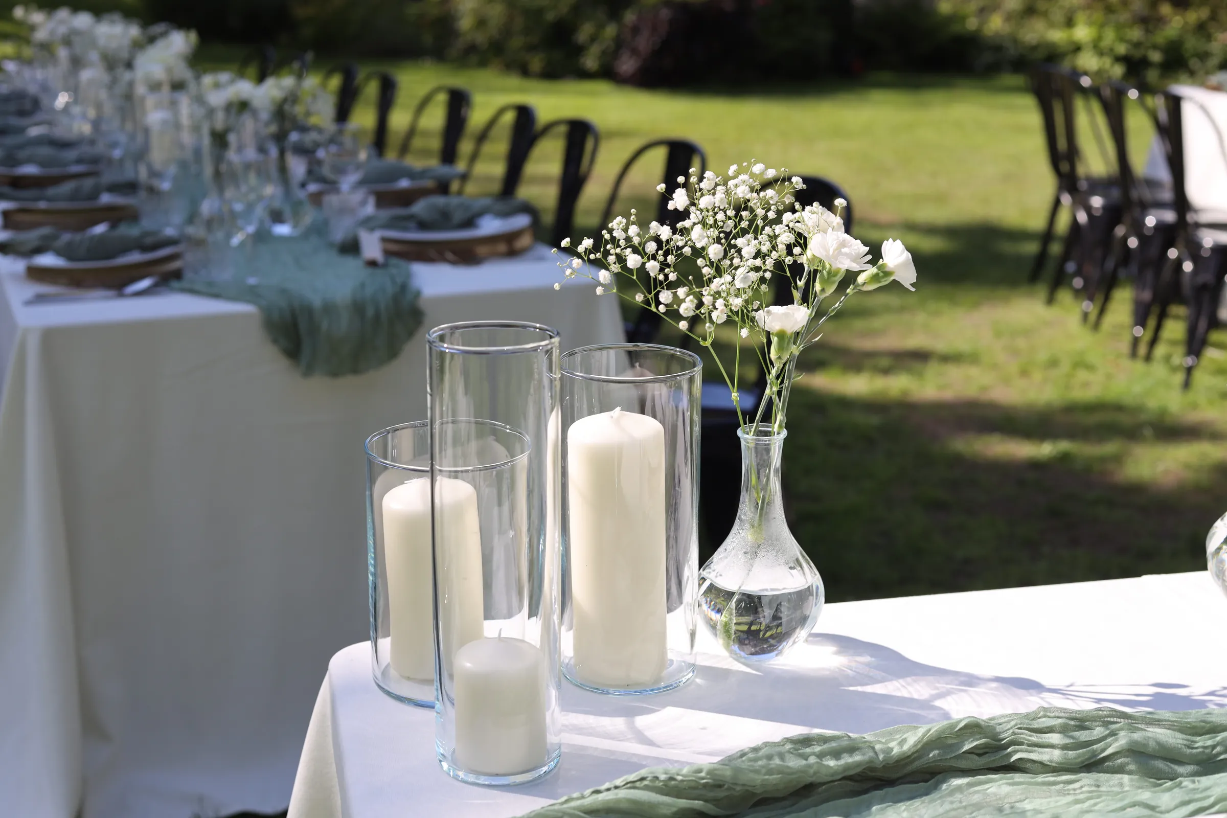White pillar candles in glass cylinders and a carafe vase with carnations and gypsophila on a table with a sage gauze table runner