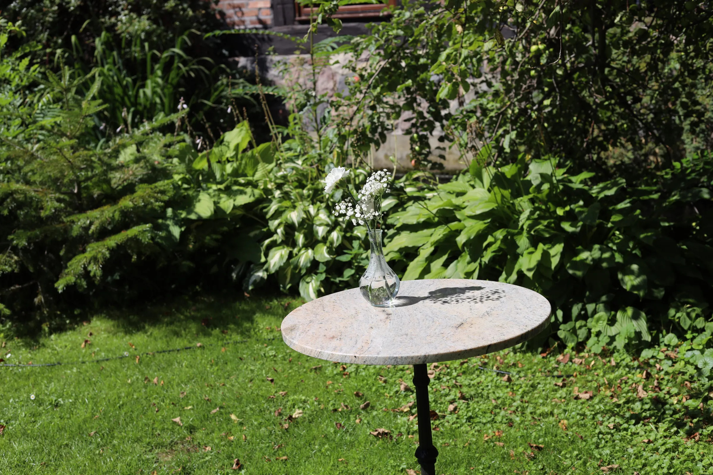 A round stone table in the garden with a carafe vase in which there is a white carnation with gypsophila, hostas and a lawn around it