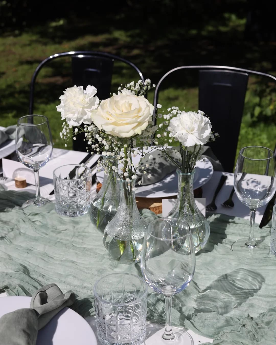Three glass vases with a white rose, carnations and a gypsophila on a sage table runner, next to them there are wine glasses and tumblers