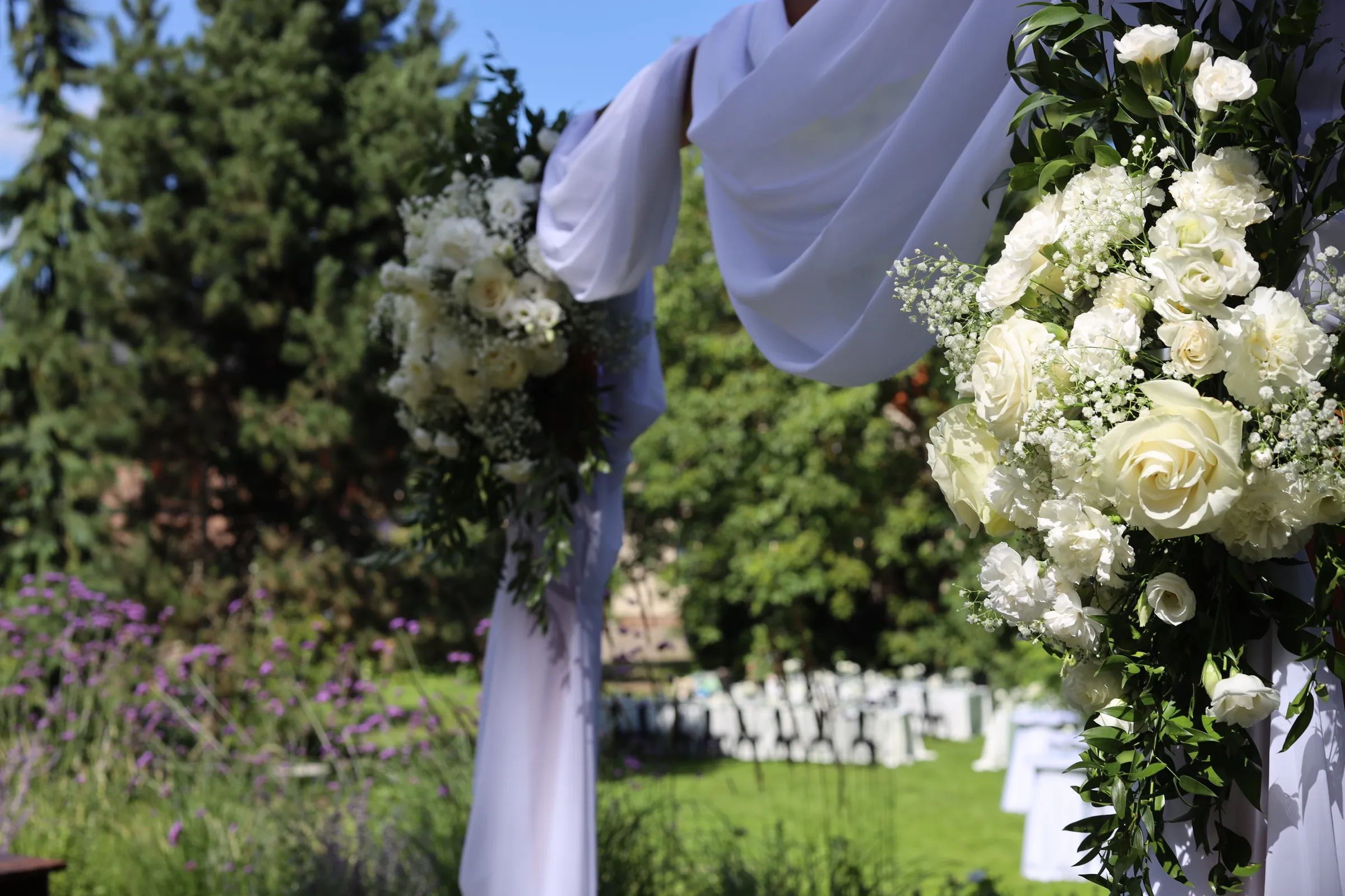 Corner compositions of white roses, carnations and gypsophila on an arch with white drapery, in the depths of the garden, white tables on the lawn