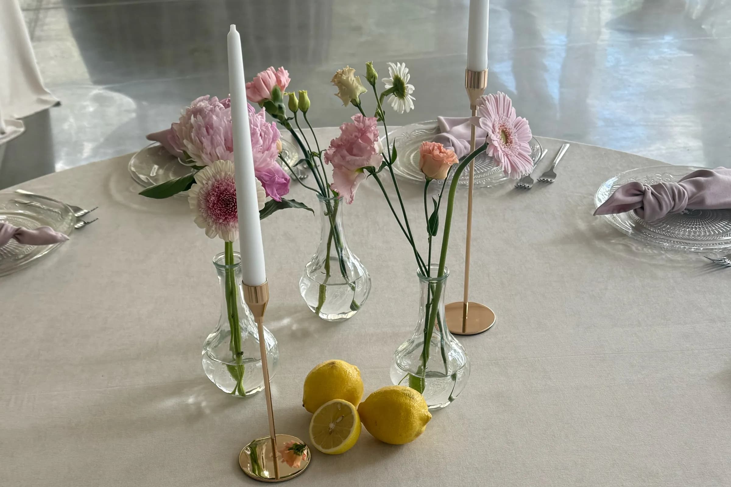 Glass vases with single flowers, whole and cut lemons and candles in gold candlesticks on the guests' table