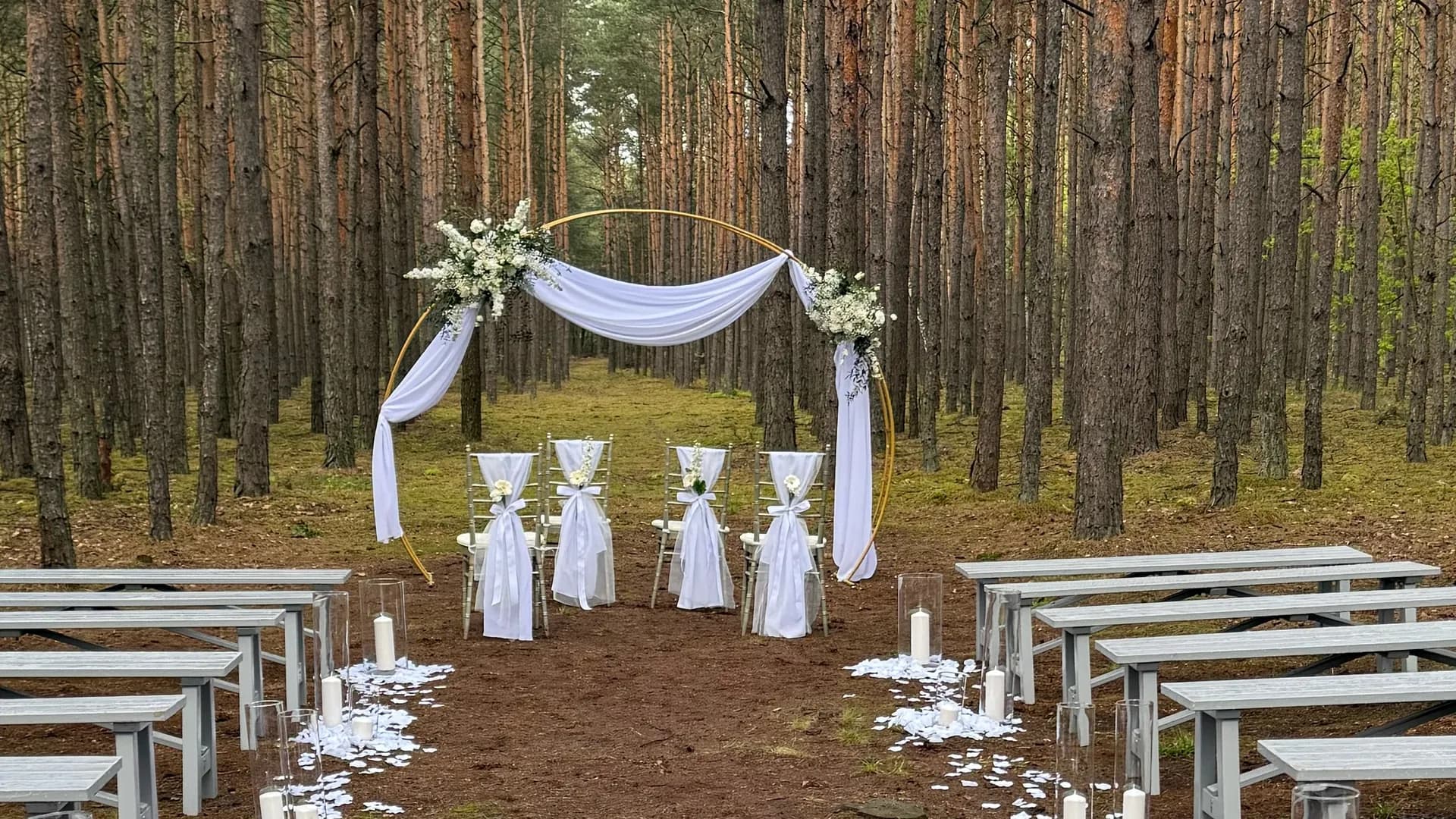 A golden round arch with white drapery and white flower arrangements, in front of it chiffon chairs with bows