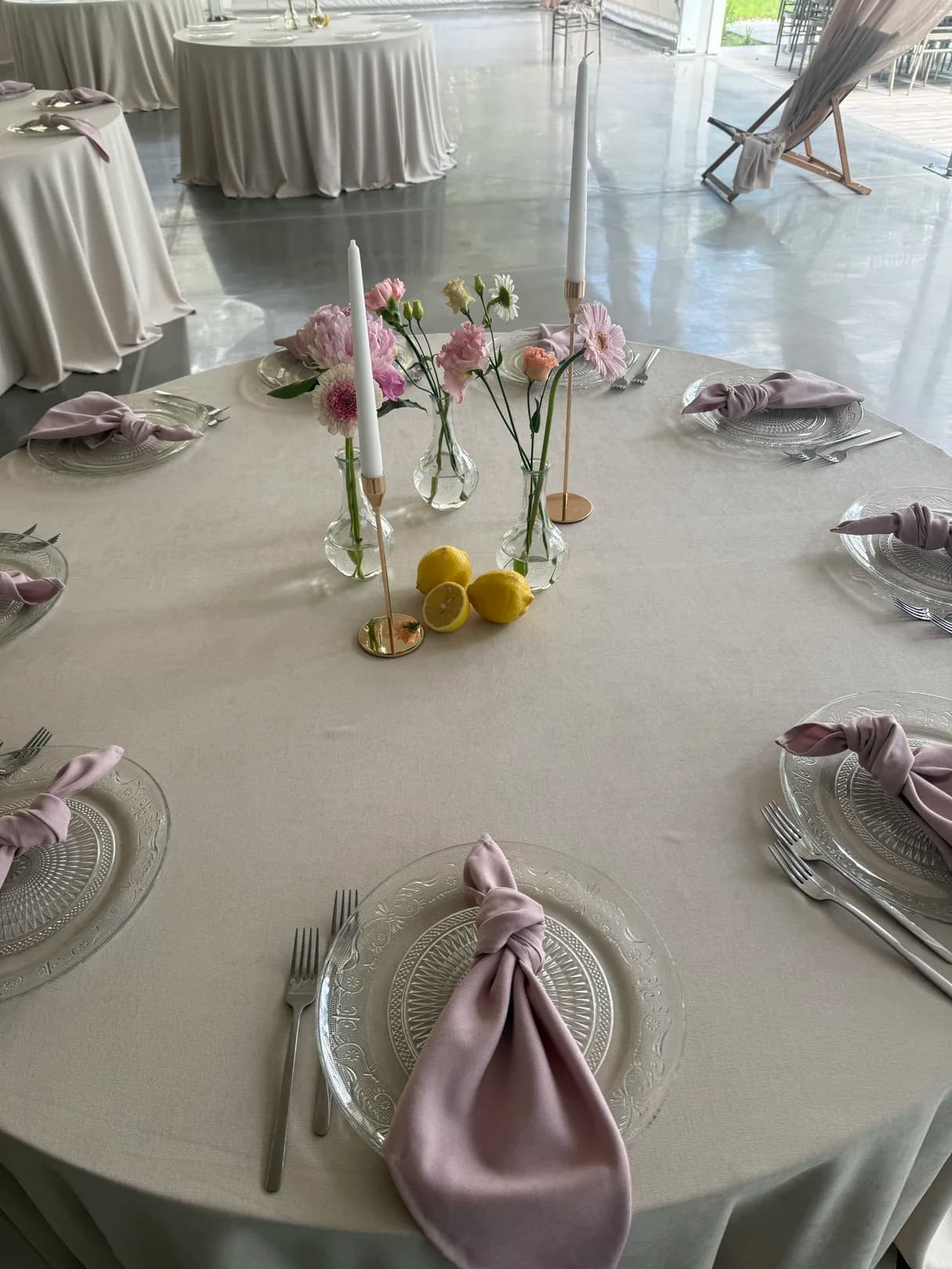 Round guests table with glass charger plates, heather satin napkins, vases with pink flowers, candles and lemons