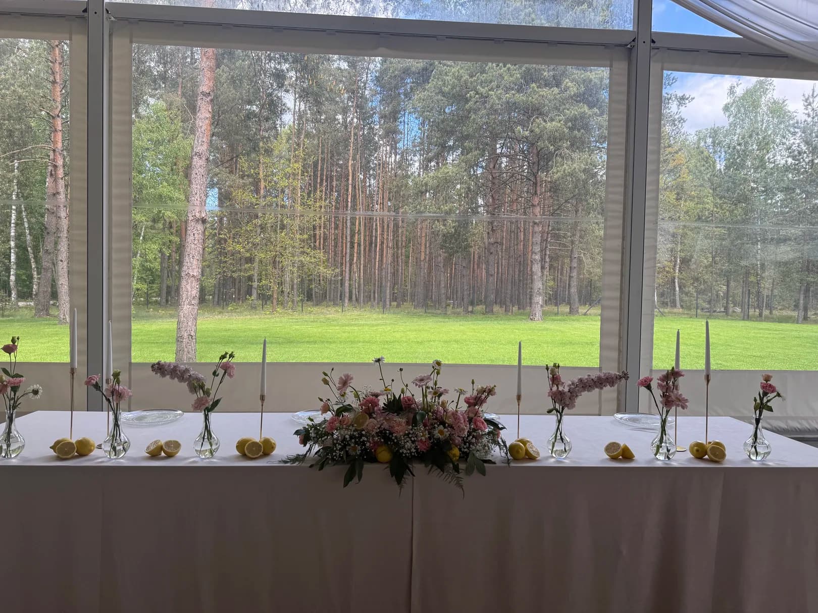 A couple's table in a glass pavilion overlooking the forest: a low composition of pink flowers, vases, candles in gold candlesticks and lemons