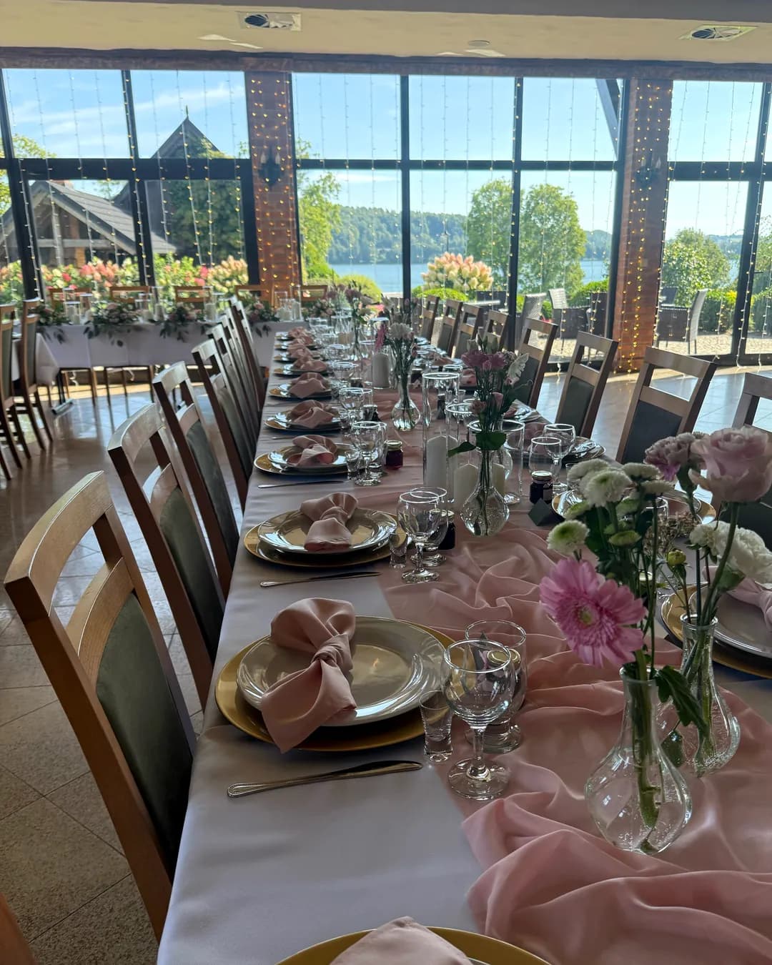 A long table with a pink table runner and low vases with gerberas, a glass wall with a view of the lake in the background