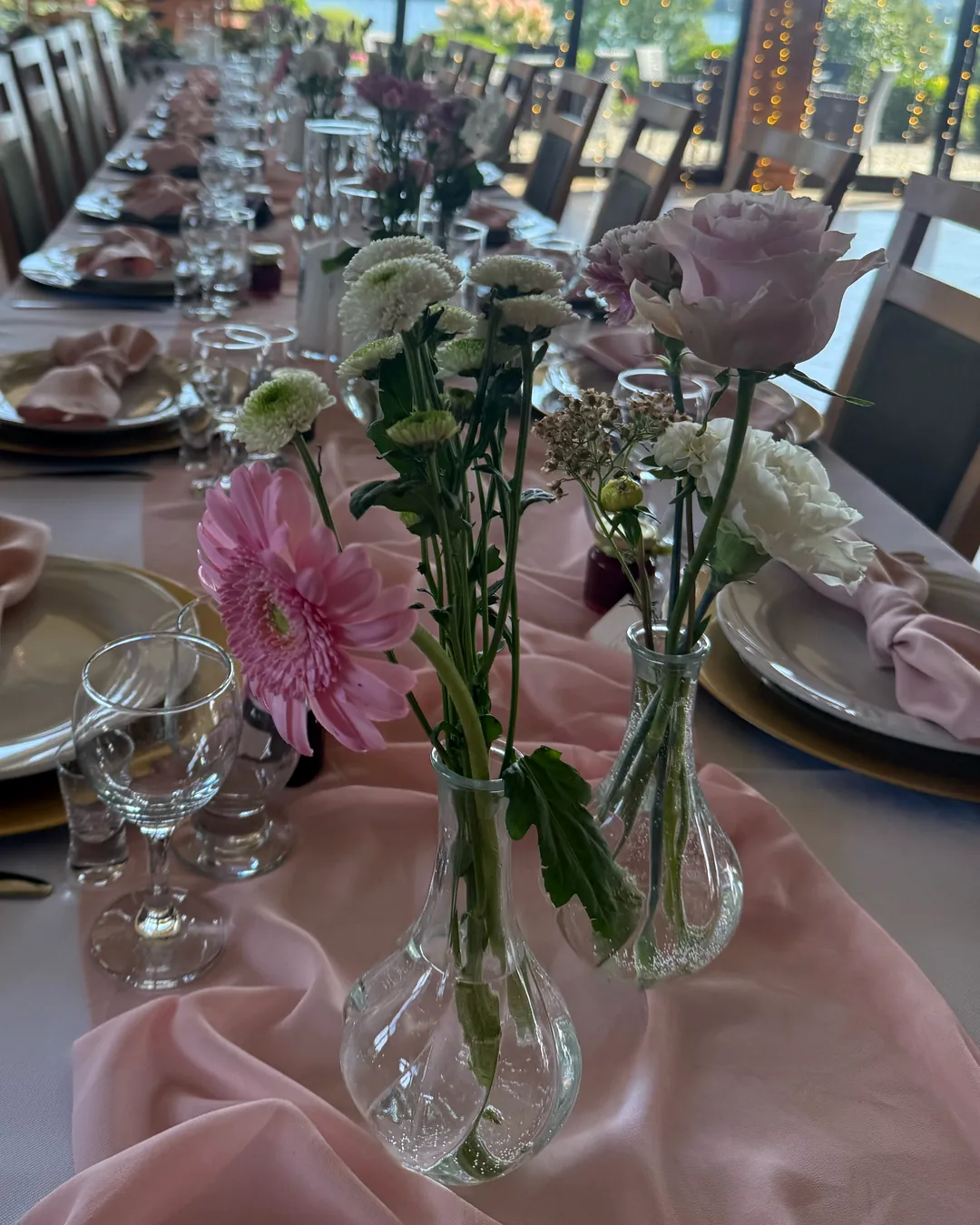 Pink gerbera, white chrysanthemums, rose and carnation in low glass vases on a pink table runner