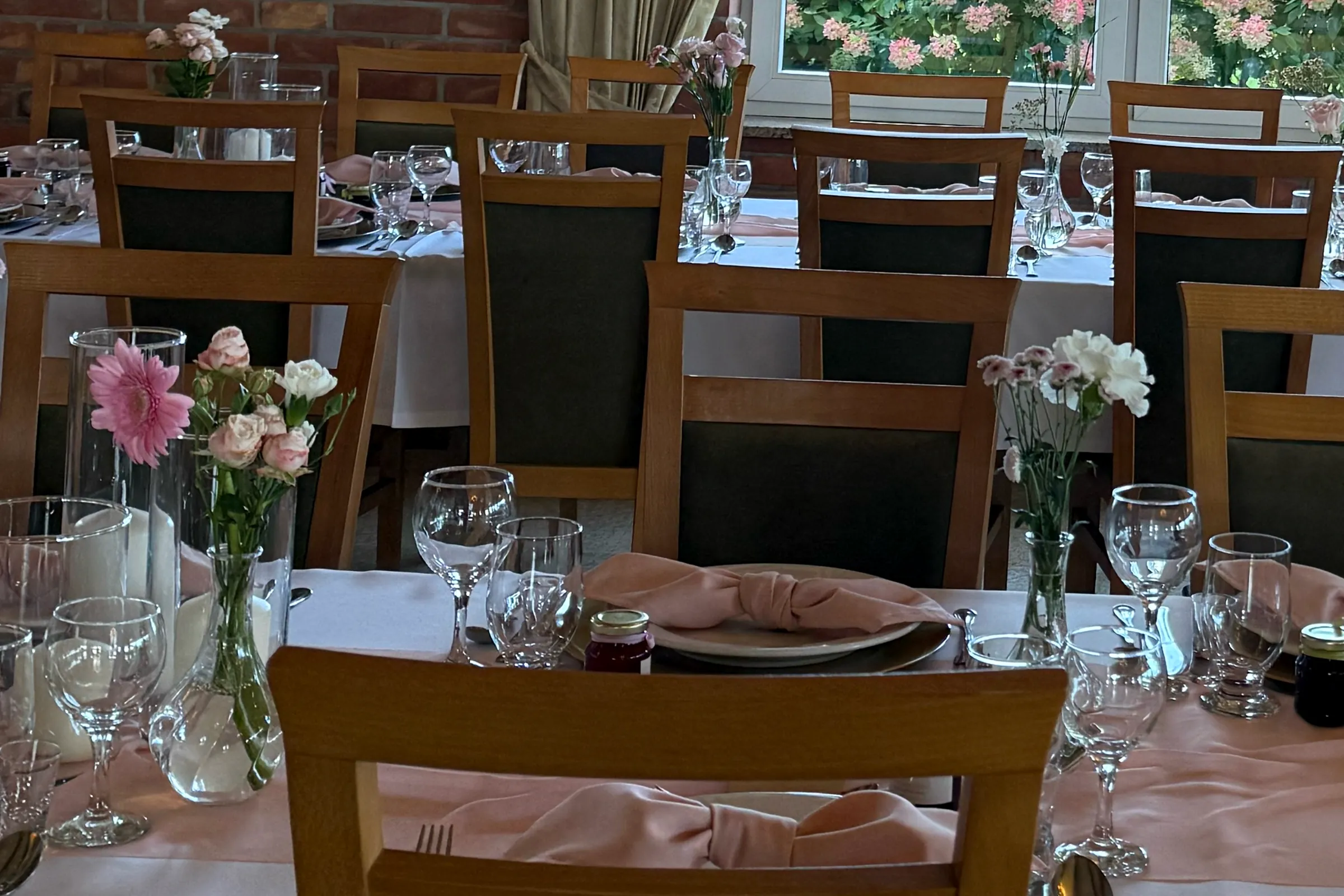 Wooden chairs, white tablecloths and pink table runners, glass vases with gerberas, roses and carnations on the tables.
