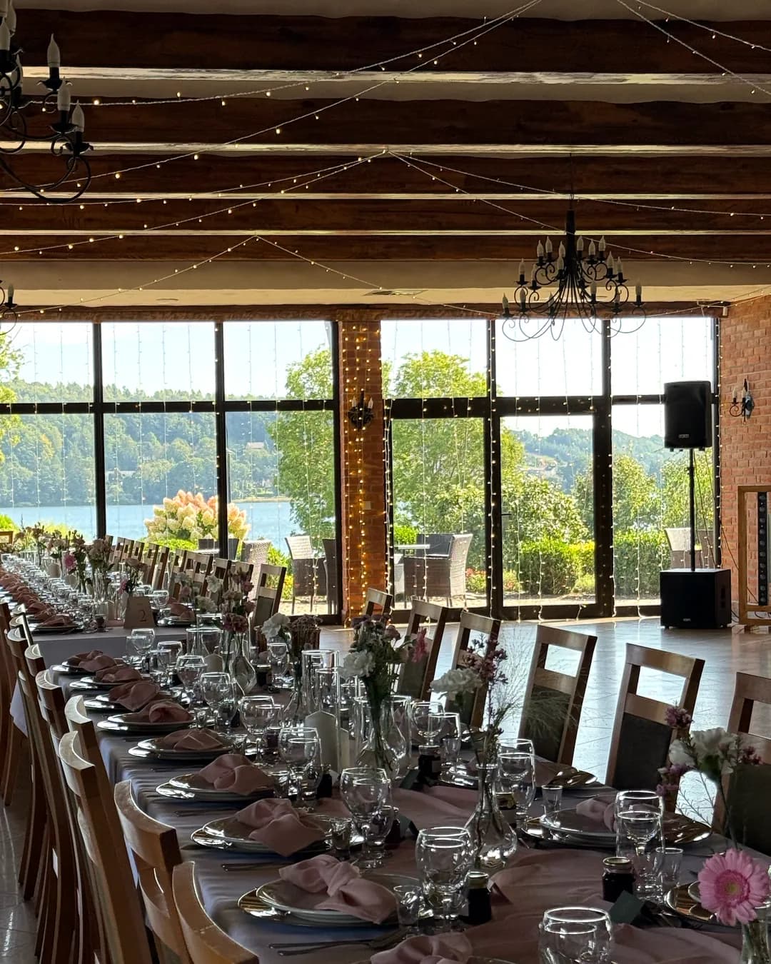 A room with wooden beams, chandeliers and garlands of lights, long tables leading to windows overlooking the lake.