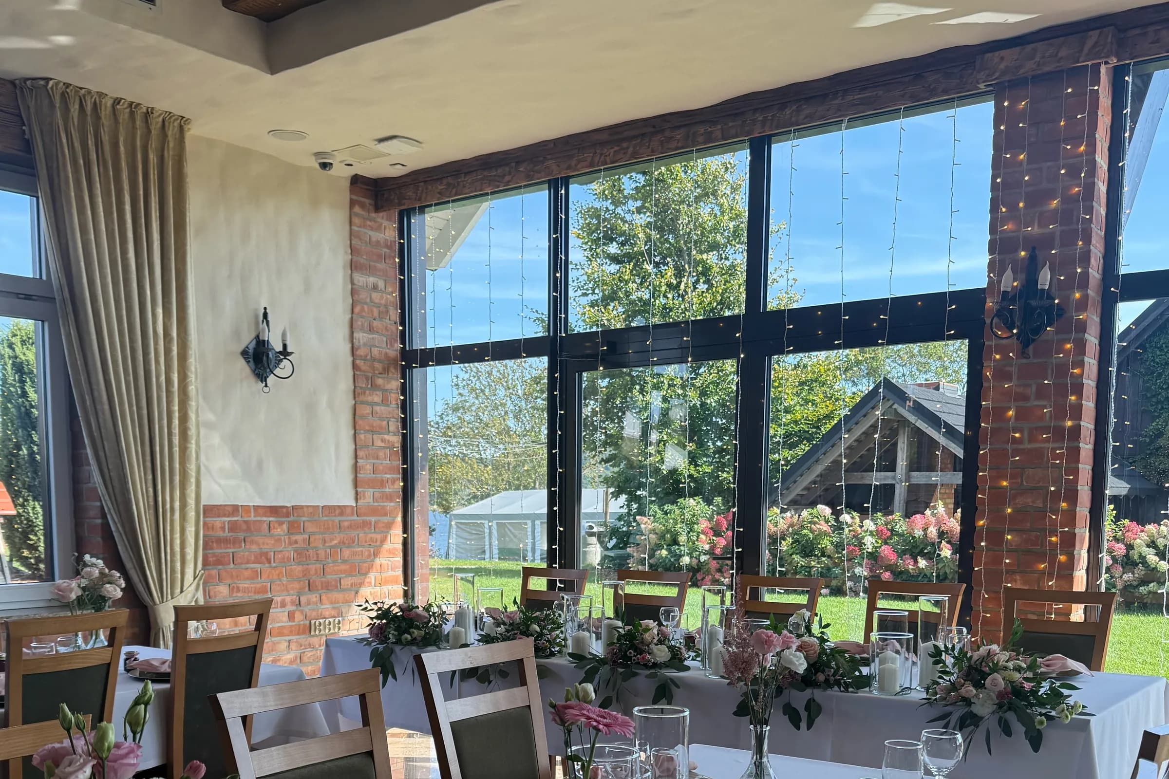 The bride and groom's table near a glass wall with a curtain of lights, a wooden beam, brick and pink flowers on the tables