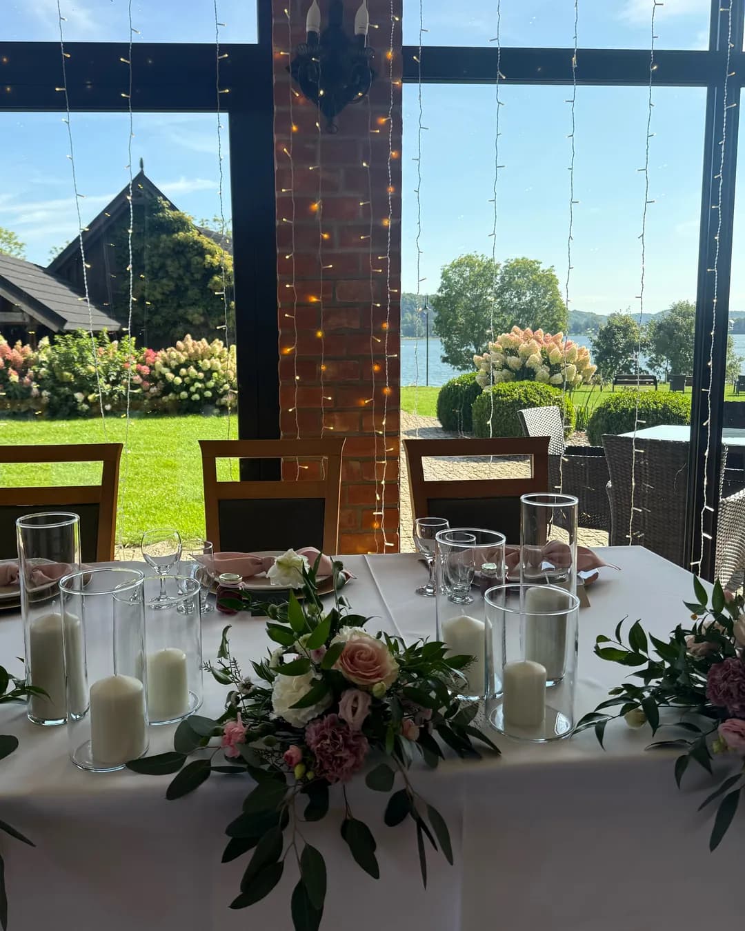 Table of the bride and groom with a garland of roses, carnations and eucalyptus and candles in glass, a garden and a lake outside the window