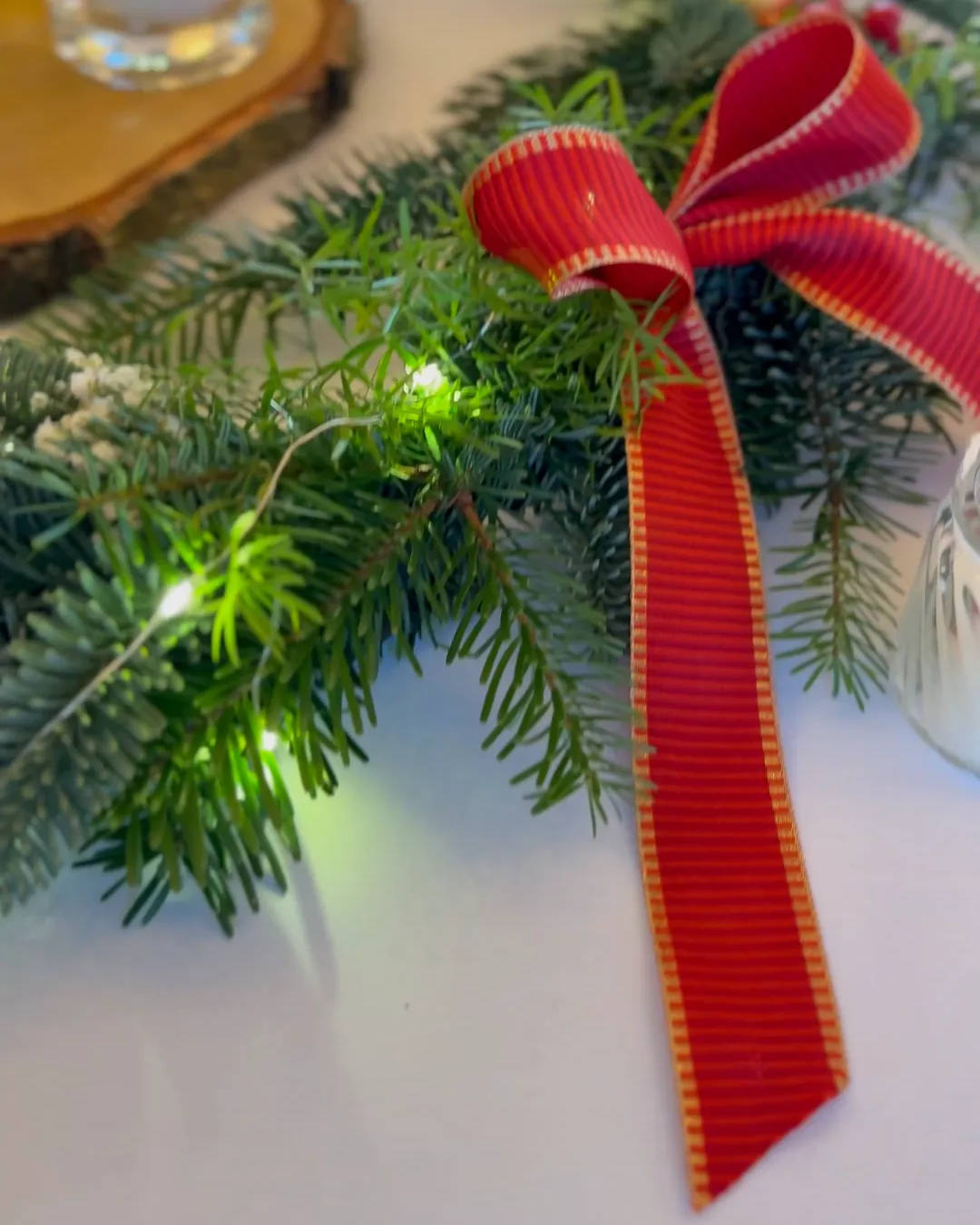 A red ribbon bow on a fir tree wreath with lights, a candle on a wood slice in the background