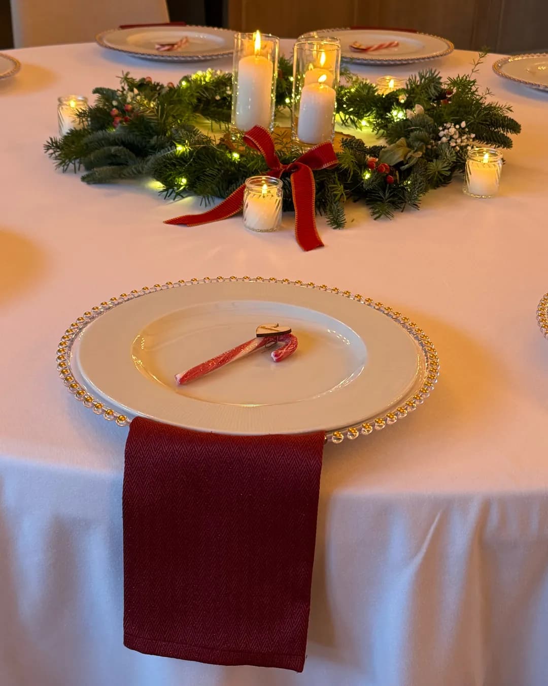 Place setting with candy cane and red napkin in front of fir wreath with candles in glass and red bow