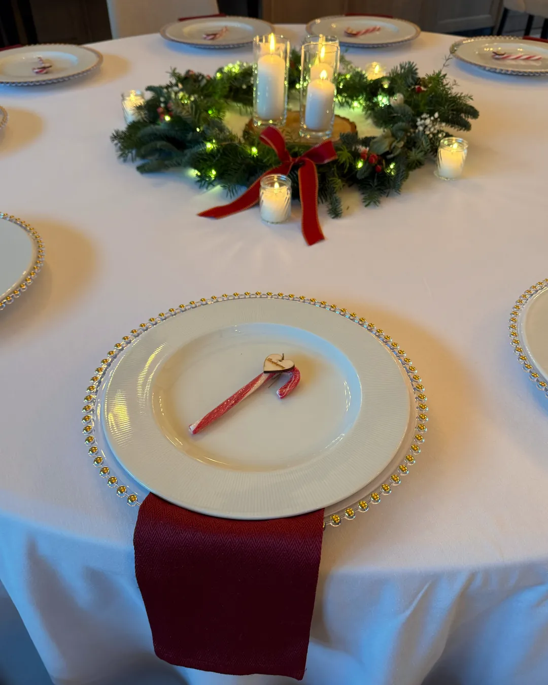 A round guests' table with a white tablecloth, plates with a gold rim, a red napkin and a fir wreath in the center
