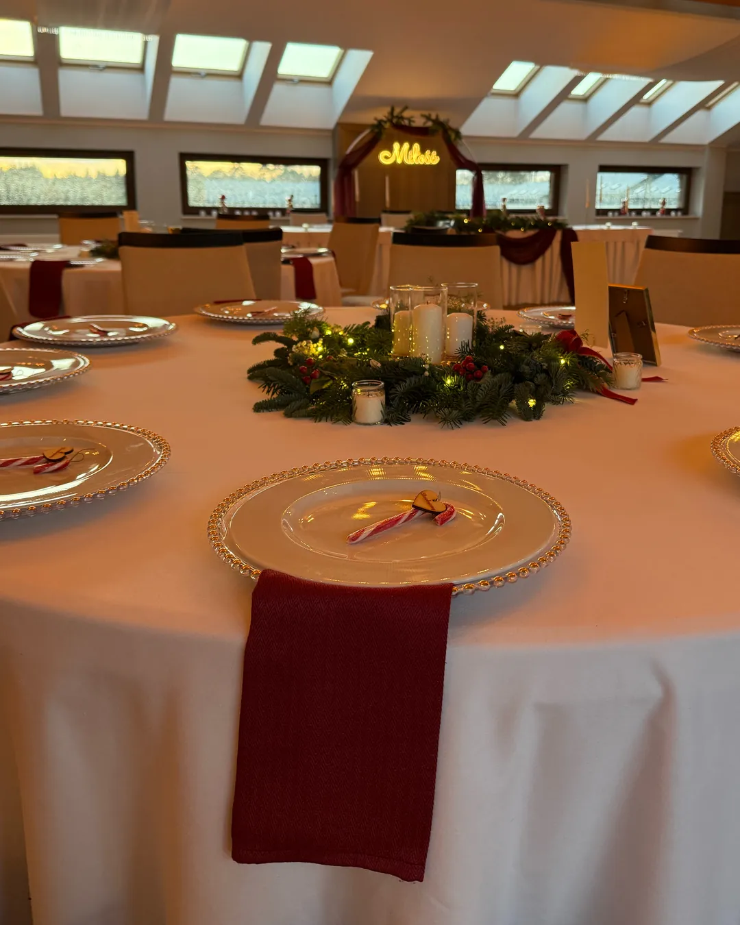 Guests' table with a plate, red napkin and fir centerpiece with candles, in the background the bride and groom's table with a neon sign