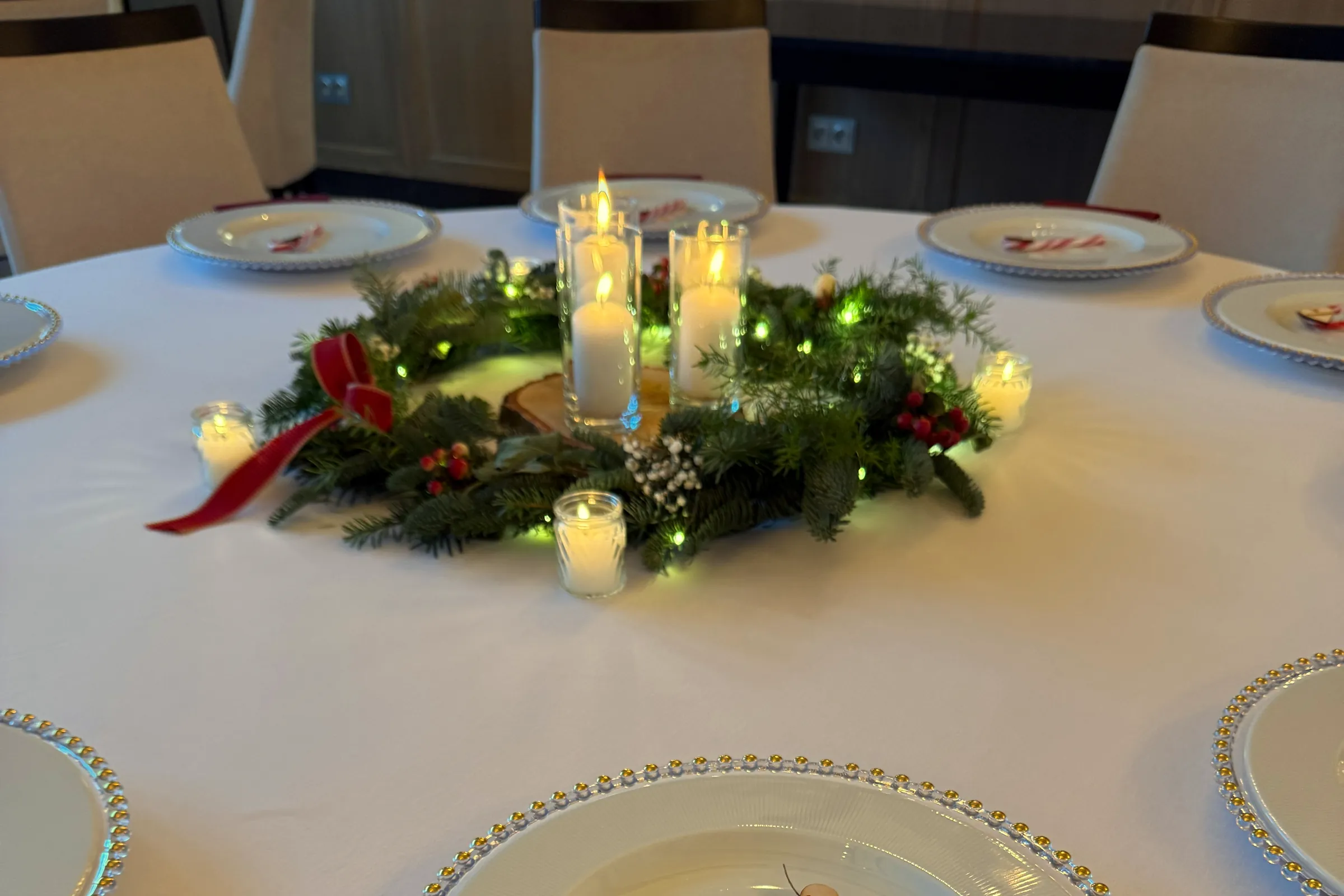 A fir centerpiece with candles in glass and a red bow in the center of a round table, plates with gold rims around