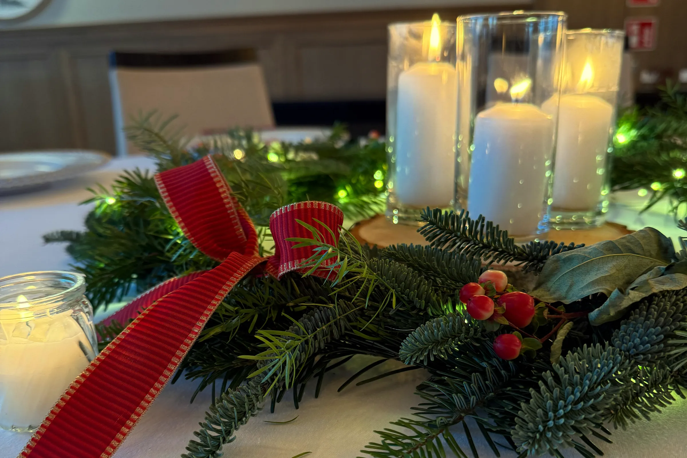 A fir tree decoration with a red bow, red fruits and three candles in a tall glass on the guests' table