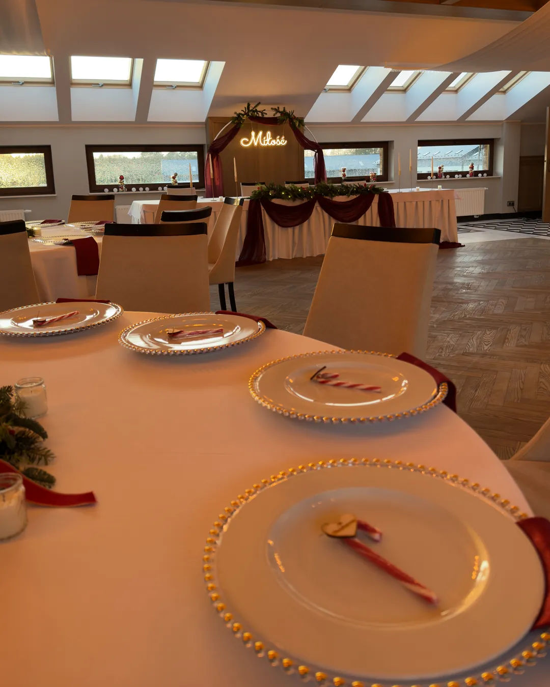 View from the guest's seat to the bride and groom's table with burgundy drapery, fir garland and a hoop with the neon sign "Love"