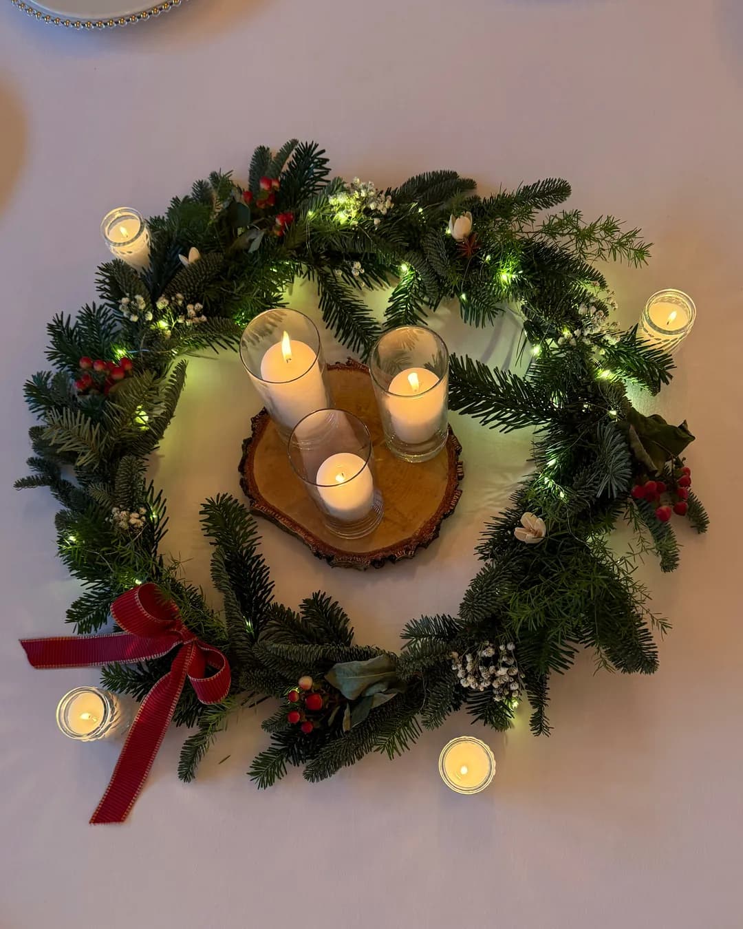 Fir wreath with lights and red bow seen from above, in the center three candles in glass on a wood slice