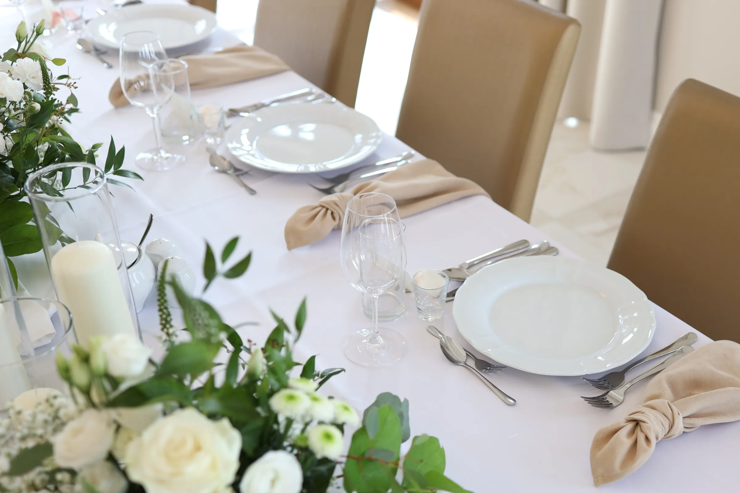 Beige cloth napkins tied in a knot next to white plates, next to them are white roses and a candle in a glass