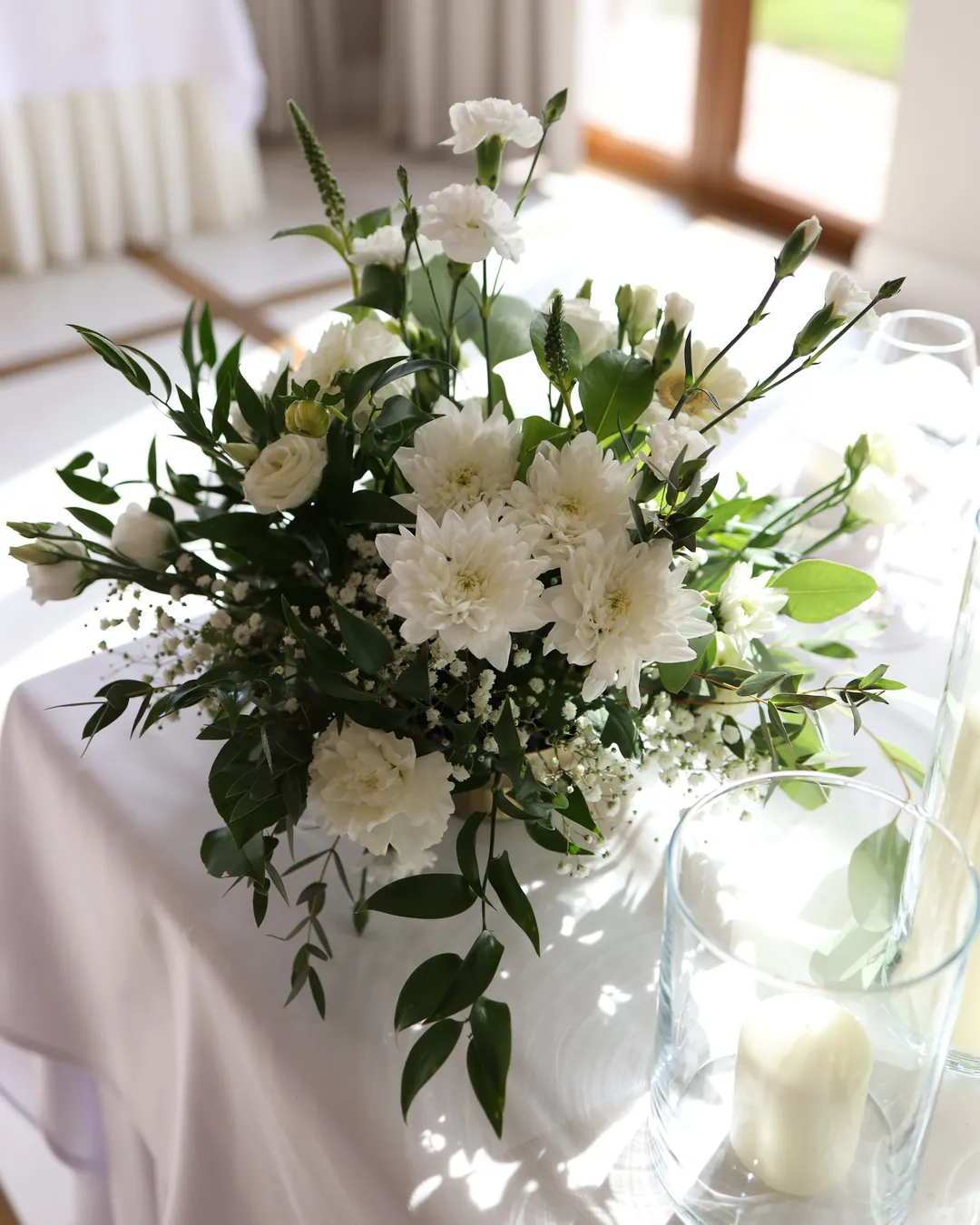 A low composition of white chrysanthemums, carnations and greenery on a white tablecloth, next to a candle in a glass cylinder