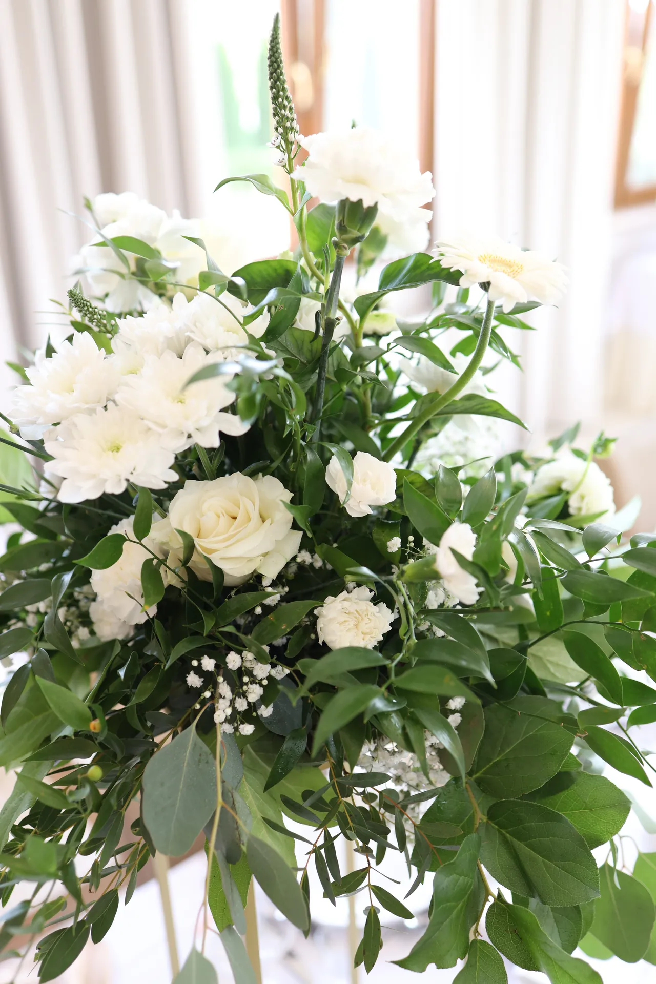 White roses and chrysanthemums with gypsophila and greenery in a composition on a tall golden flower stand, seen from close up
