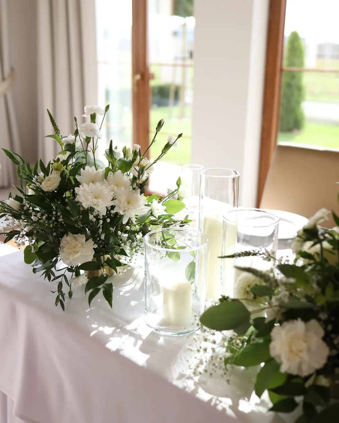 A low composition of white chrysanthemums and carnations next to candles in glass cylinders, in the sun by the window