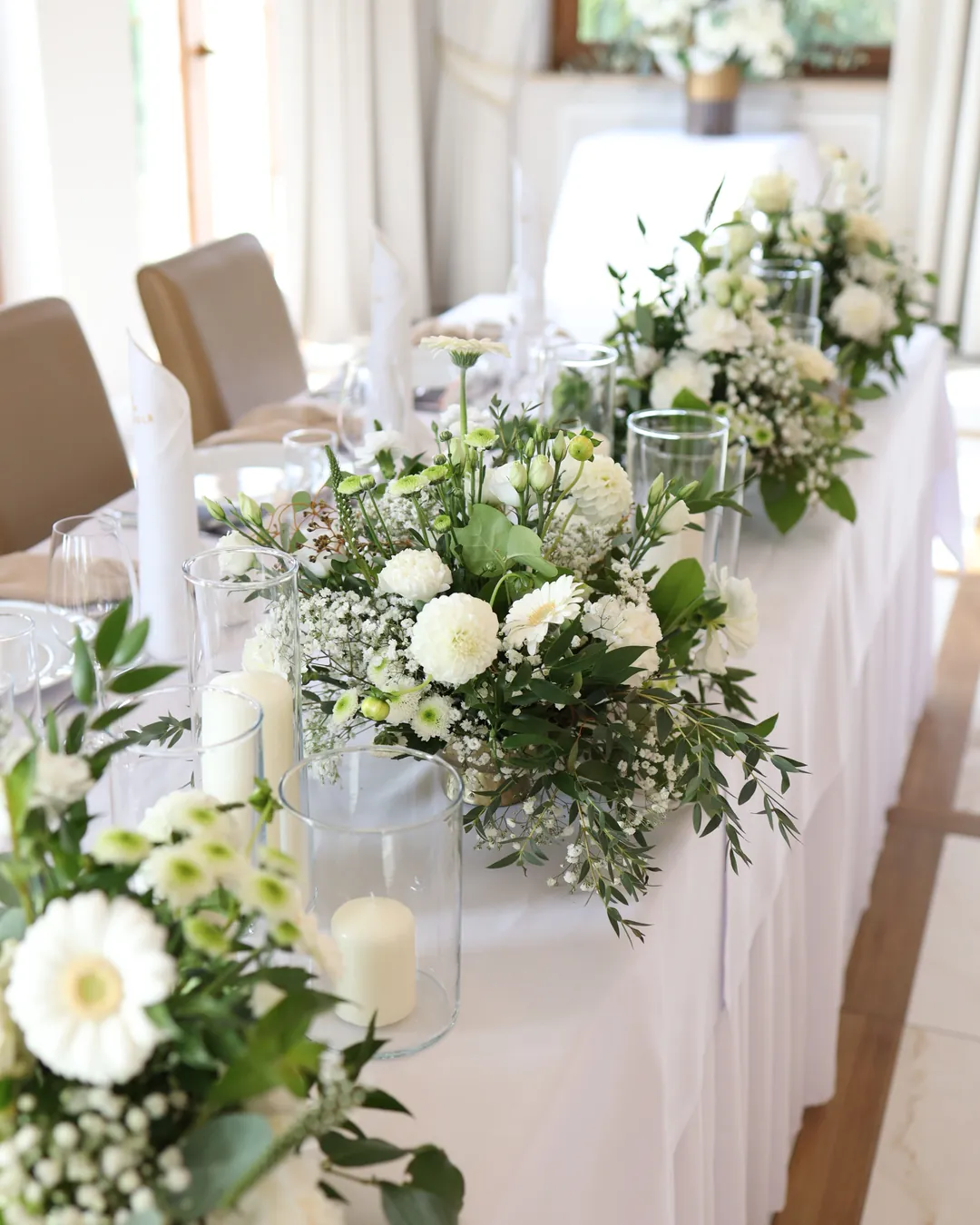 A row of low compositions of white flowers and greenery with candles in glass on the table of the bride and groom with a white tablecloth