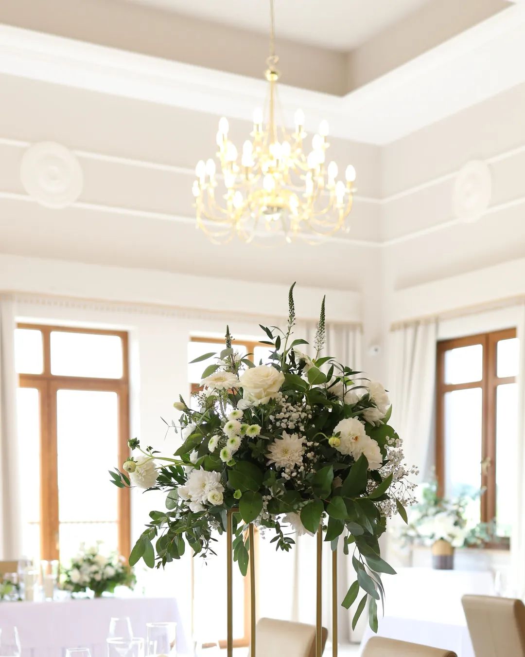 Composition of white flowers and greenery on a tall golden flower stand under a crystal chandelier in a bright hall