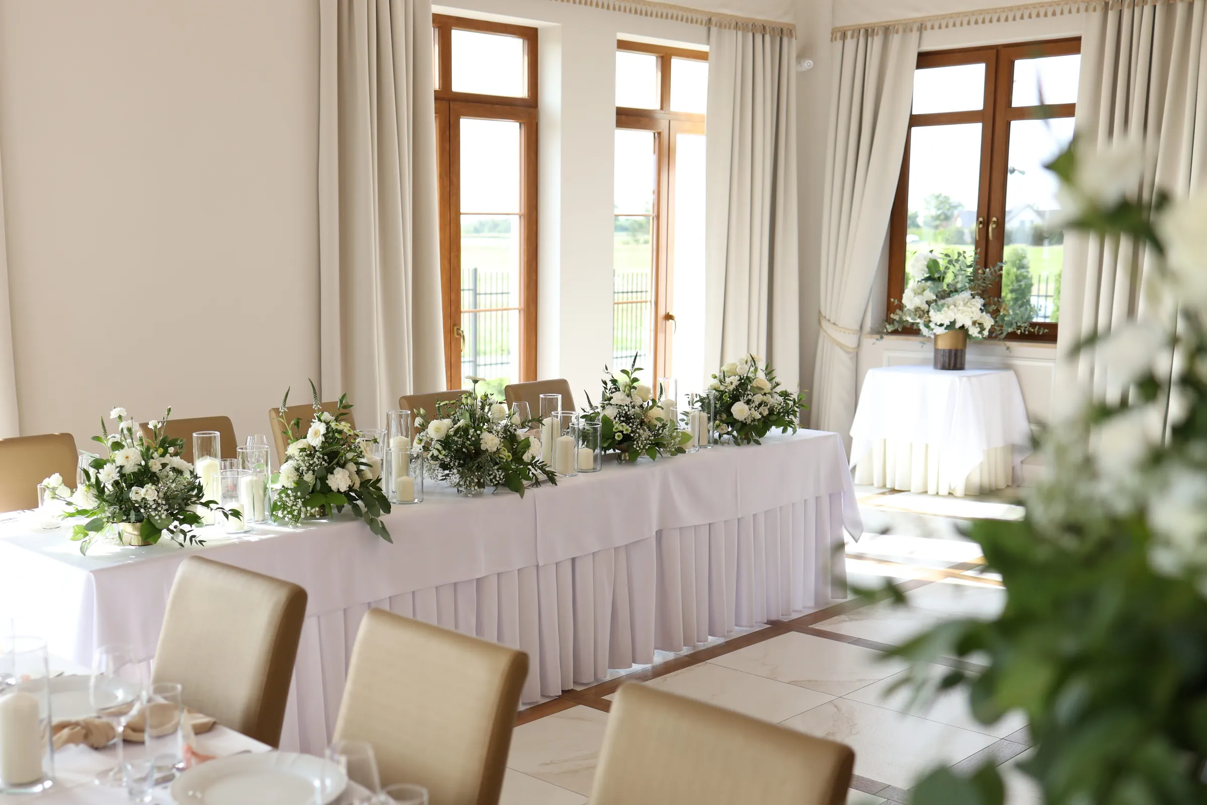 The bride and groom's table with low compositions and candles against the background of the high windows of the hall, with a table set for guests in front