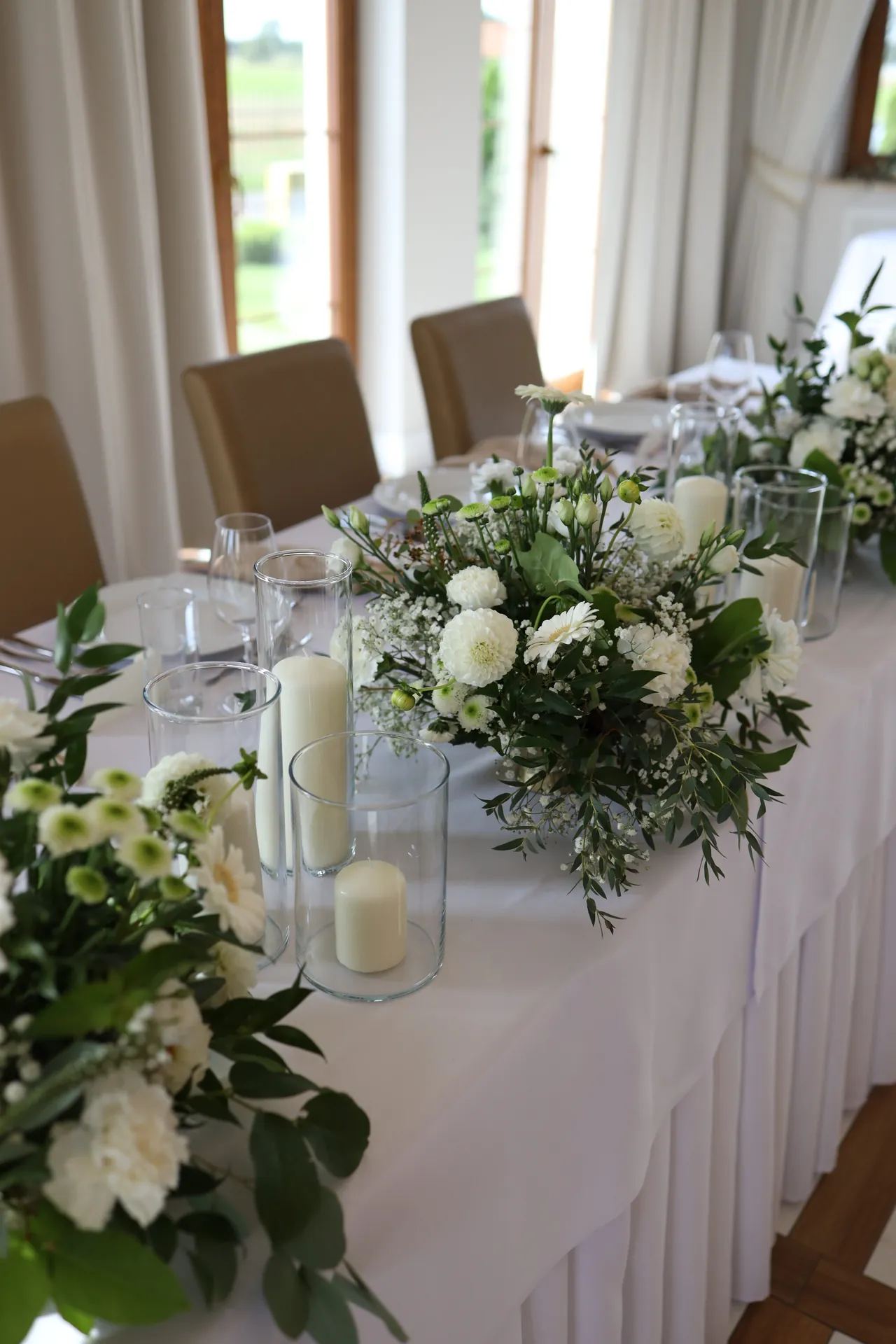 Candles in glass cylinders and low compositions of white flowers on the table of the bride and groom near the window to the garden