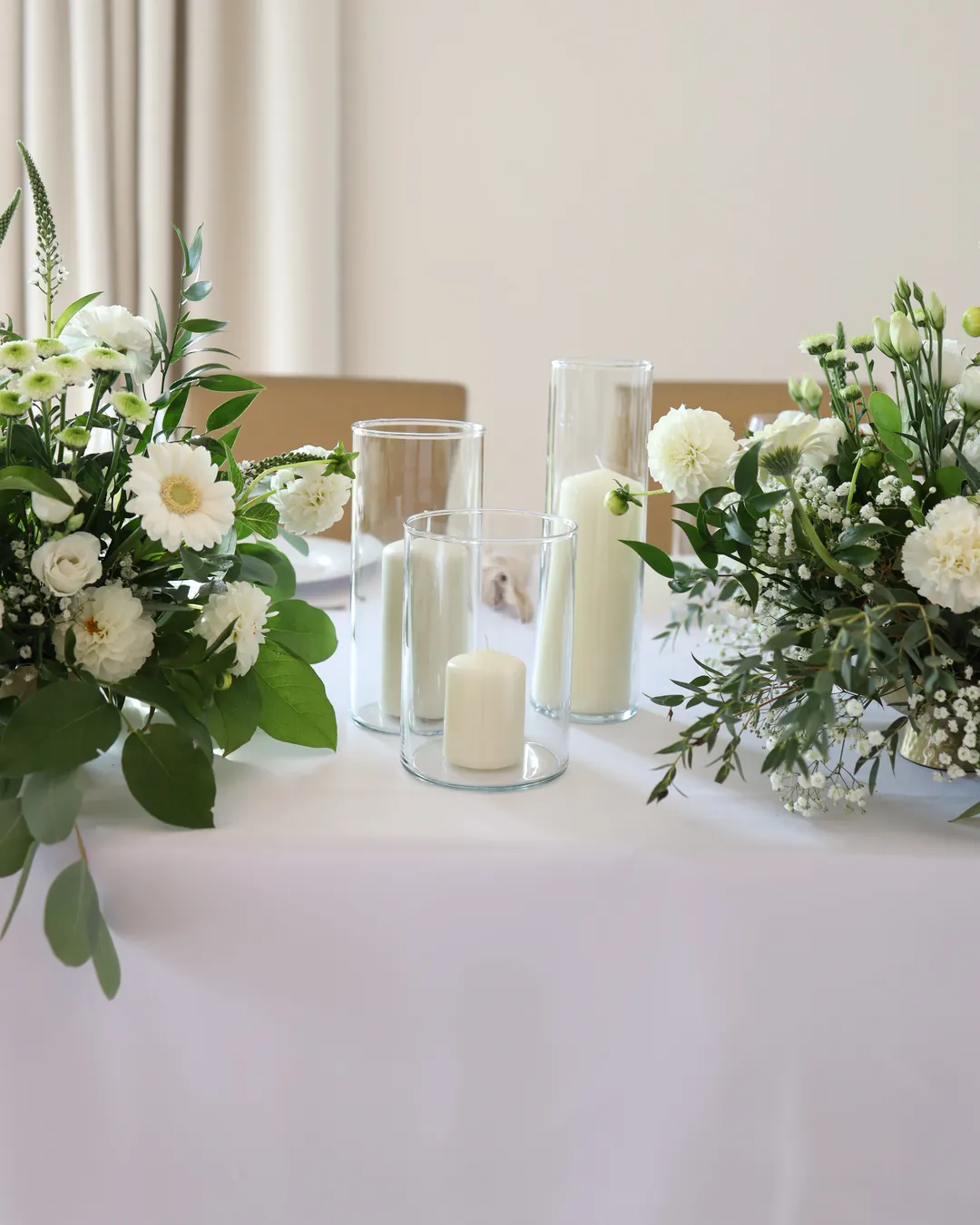White candles in glass cylinders placed between two compositions of white gerberas, roses and greenery