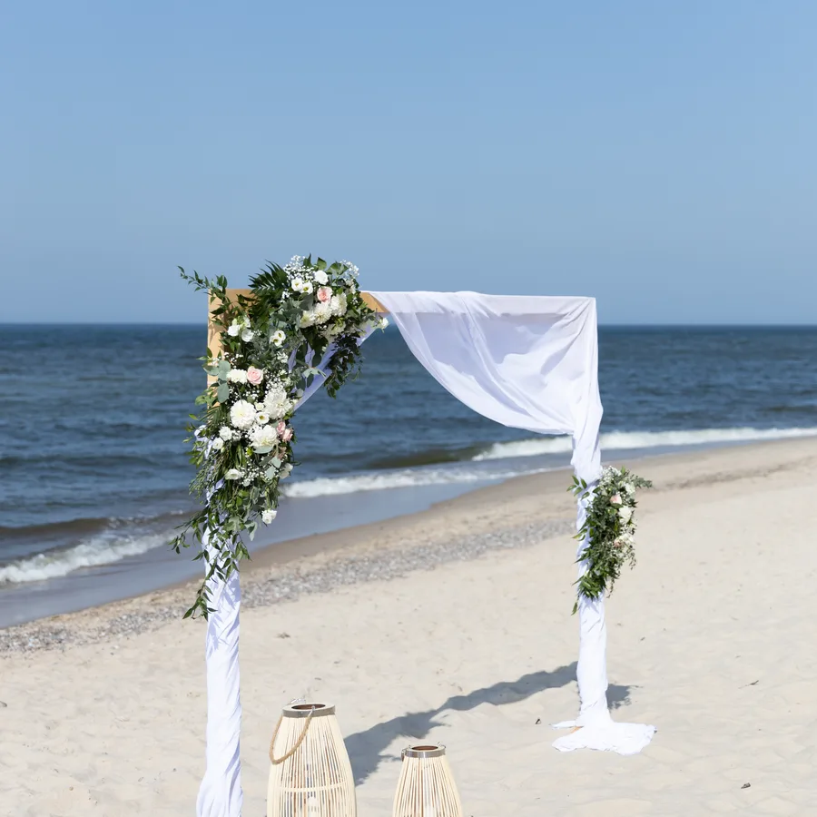 Łuk kwiatowy z białą draperią na plaży nad morzem
